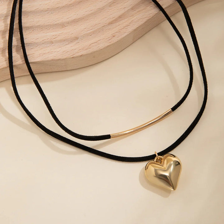 A black double-cord adjustable choker necklace featuring a curved gold bar on one strand and a shiny vintage heart pendant on the other, displayed against a light beige surface with a wooden accent.