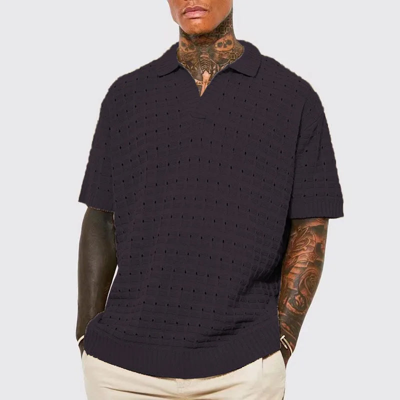 A man with tattooed arms and neck wears a dark, breathable knit design short-sleeve polo shirt and light beige pants, posing with his hands in his pockets against a light gray background.