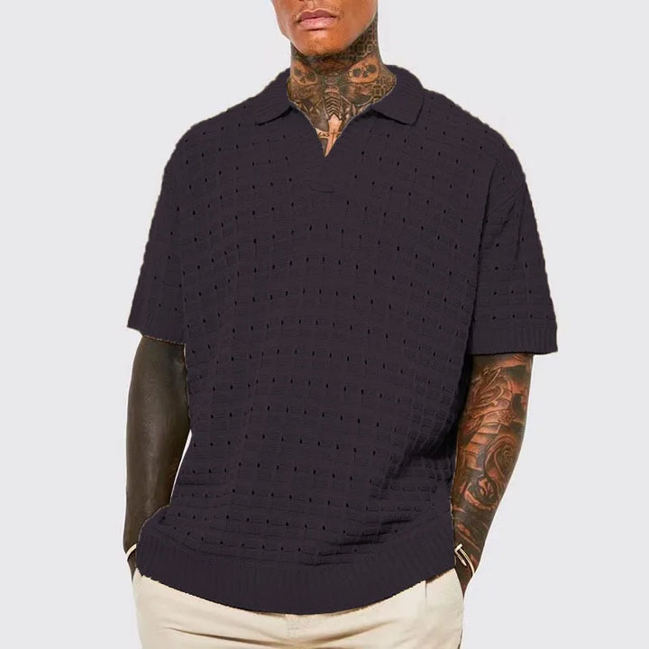 A man with tattooed arms and neck wears a dark, breathable knit design short-sleeve polo shirt and light beige pants, posing with his hands in his pockets against a light gray background.
