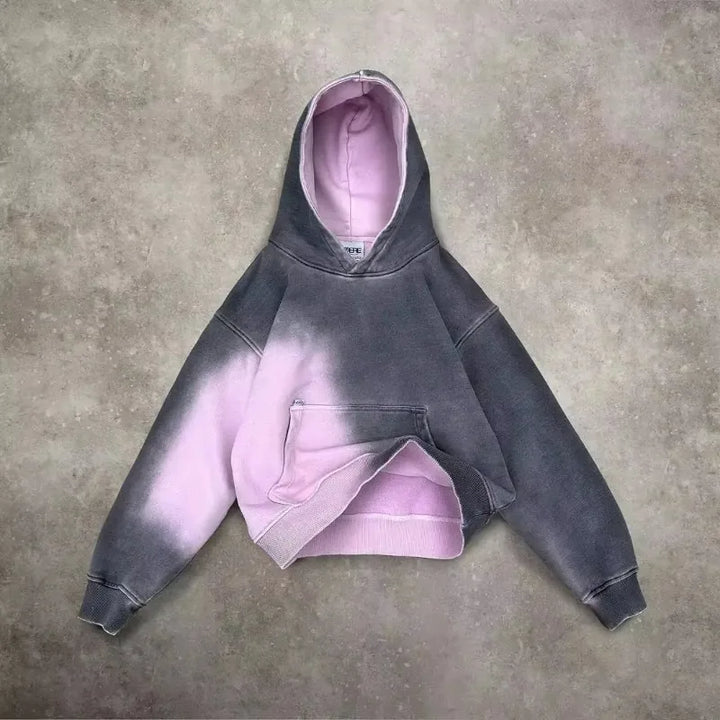 A gray and pink tie-dye hoodie with a gradient design, front pocket, and hood is laid flat on a textured gray surface. The inside is light pink, contrasting with the darker exterior for a unique look.