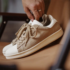 A hand holds a Lowland Odyssey Sneaker, crafted with beige suede and white accents, resting on a light brown chair. Another matching minimalist sneaker sits next to it.