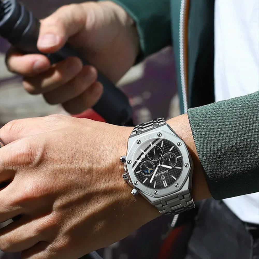 A person wearing a stainless steel watch with chronograph dials and a black dial is holding a black object, possibly a microphone, with both hands. The green jacket over a white shirt adds to the smart look.