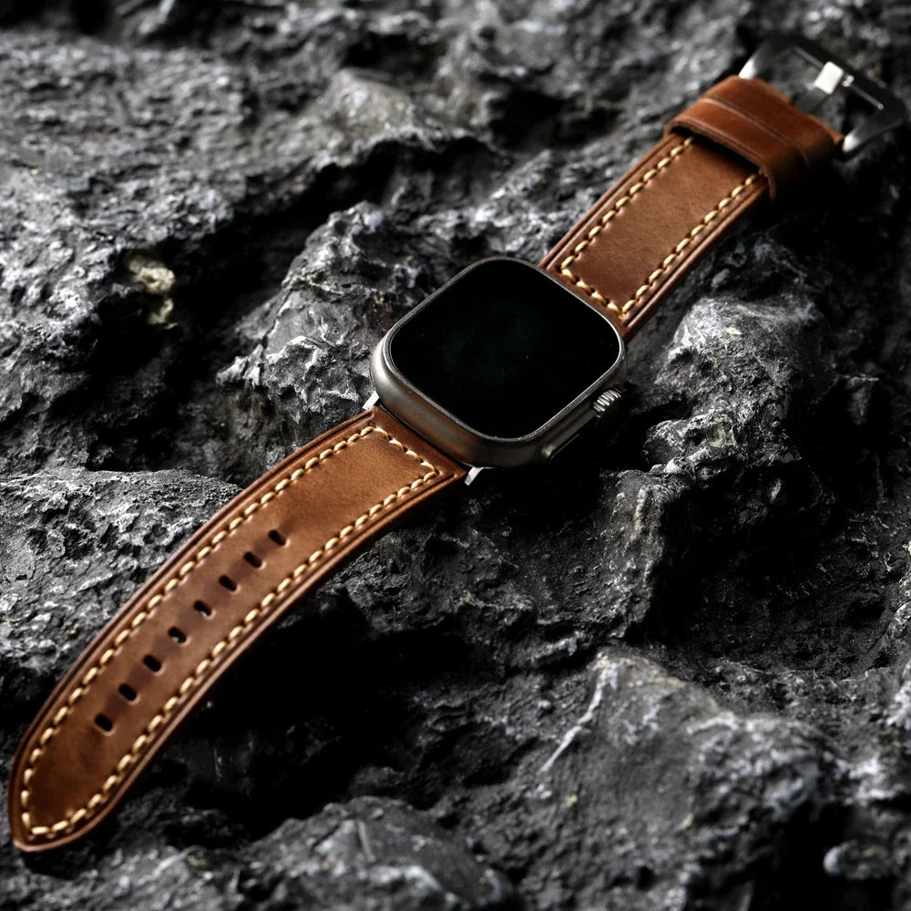 A smartwatch with a handcrafted cowhide leather strap and white stitching is placed on a textured, dark gray rocky surface.