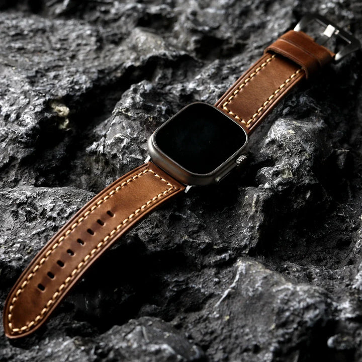 A smartwatch with a handcrafted cowhide leather strap and white stitching is placed on a textured, dark gray rocky surface.