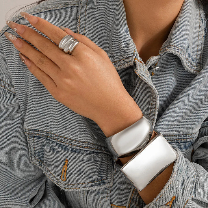 A person wearing a light denim jacket displays their hand adorned with a chunky, punk-inspired silver ring and two bold geometric design bracelets—unisex jewelry that stands out against long, nude-polished nails.