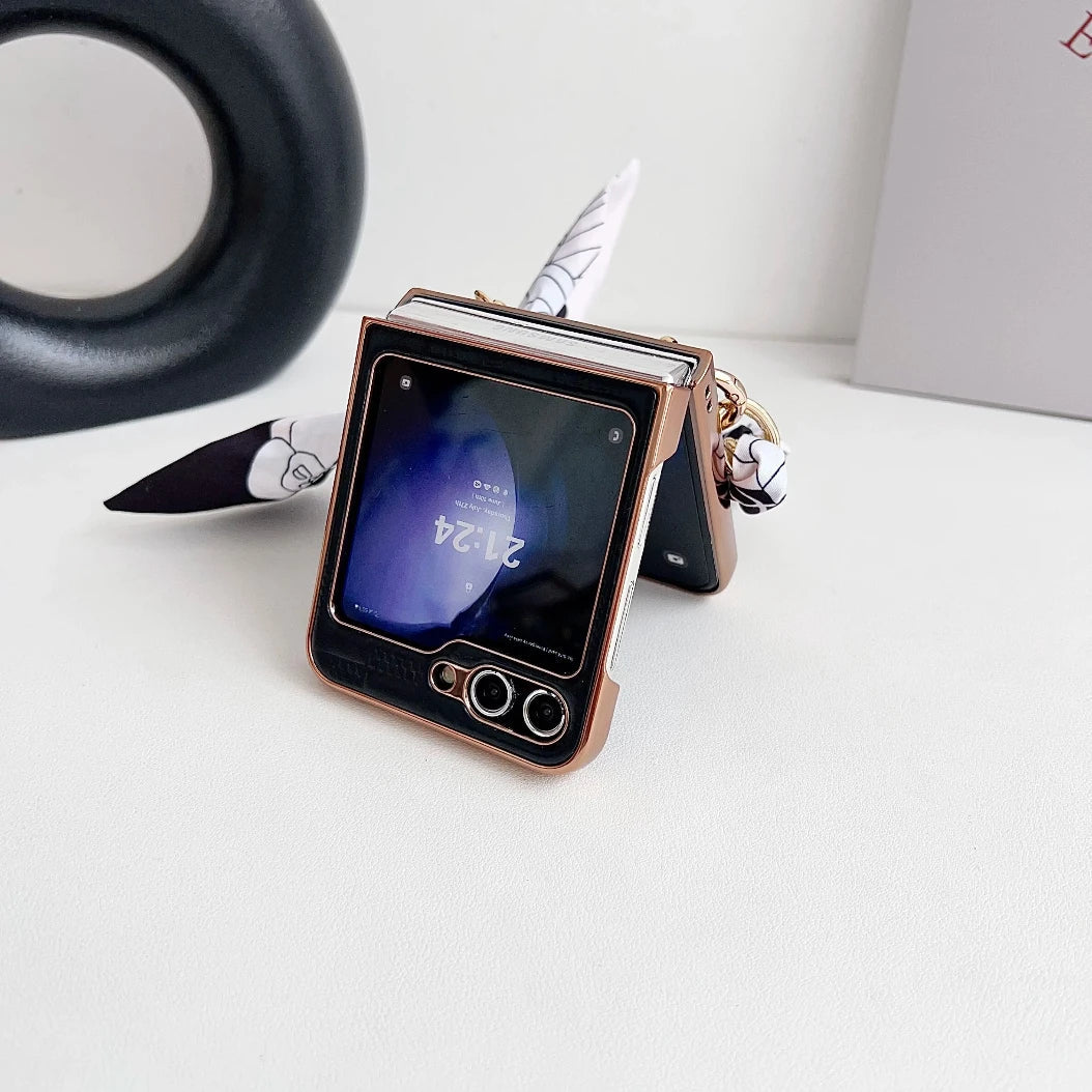 A Samsung Galaxy Z Flip in a premium leather case is partially open and standing on a white surface. A patterned ribbon is attached for style, while the screen displays the time as 21:24, offering both elegance and shockproof protection.