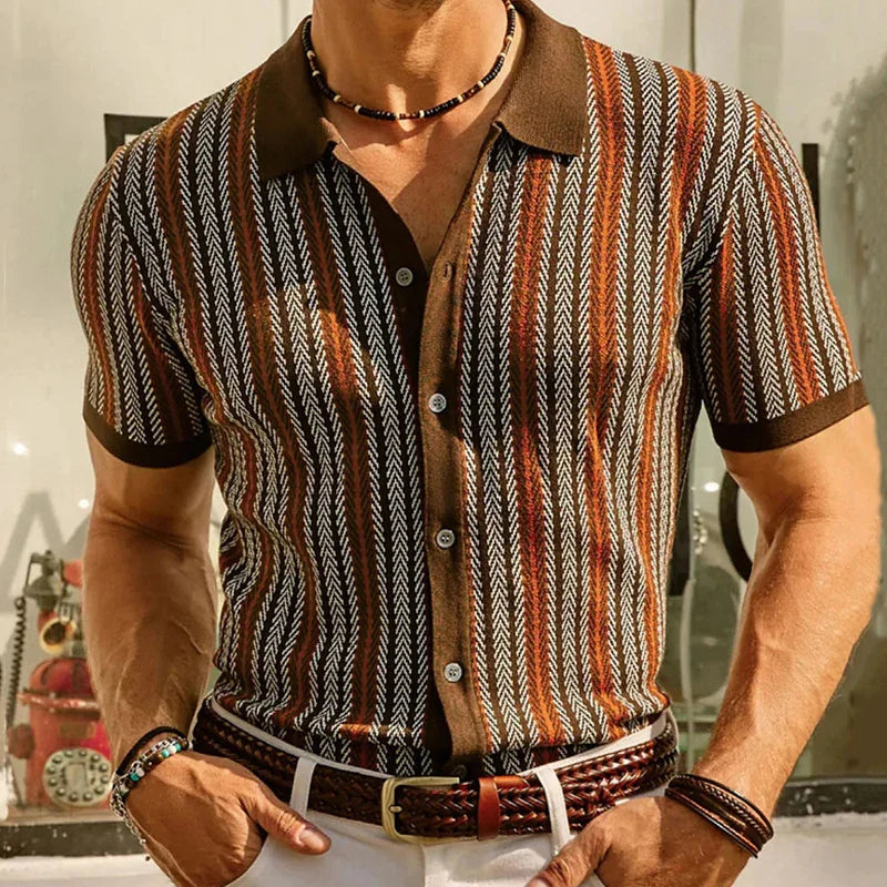 A man wearing a striped, short-sleeve button-up shirt crafted from a linen cotton blend in brown, orange, and white tones with a dark collar. He accessorizes with a beaded necklace, bracelets, a brown belt, and white pants. His hands are in his pockets.