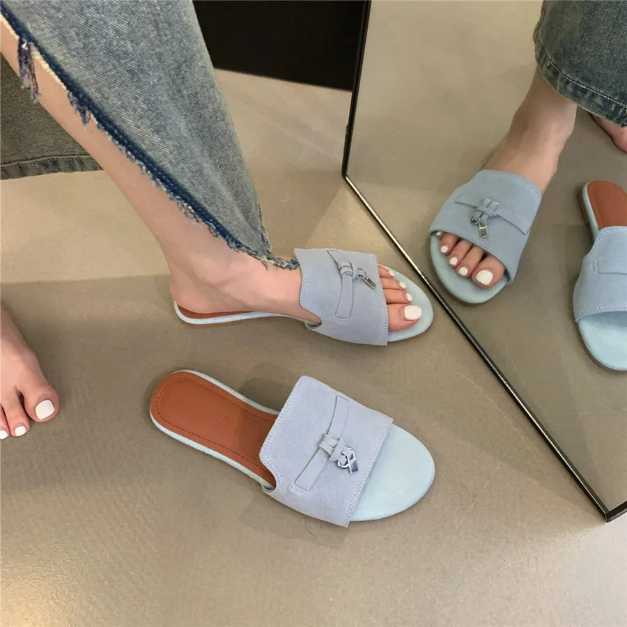 A person wearing light blue slide sandals with a decorative buckle and cushioned insole stands near a mirror, showing both feet and the reflection. The person is dressed in frayed-hem jeans and has white-painted toenails.