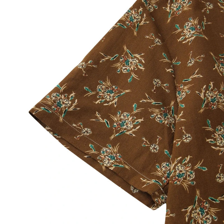 Close-up of a brown short-sleeve cotton shirt with a floral pattern in light blue, beige, and green tones. The fabric appears soft and lightweight, featuring a relaxed fit and classic design for easy, casual style.