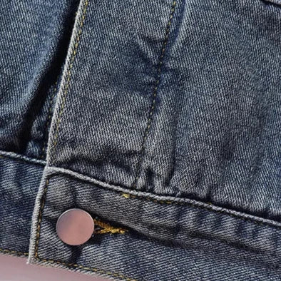 Close-up of blue denim fabric showing a seam, yellow stitching, a metal button, and a pocket flap detail. The texture of the denim weave is clearly visible, highlighting its Retro Style charm and subtle Denim Patchwork elements.
