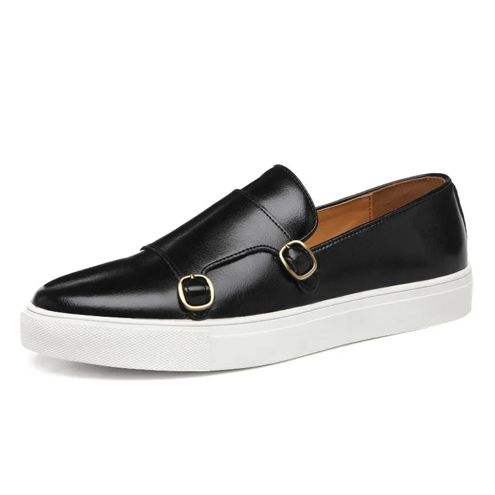 A black leather men's casual slip-on loafer with a white rubber sole and two gold buckles on the upper, viewed from the side against a white background, showcases classic British fashion elegance.