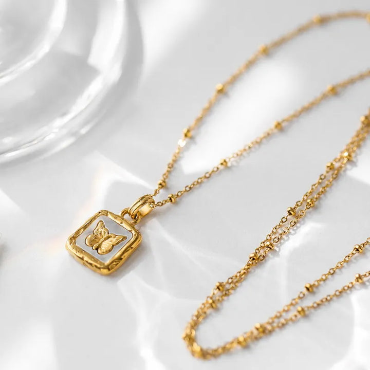 A delicate gold necklace with a square pendant featuring a raised butterfly design, showcasing elegant craftsmanship and minimalist design, displayed on a white surface with soft, natural light reflecting around it.