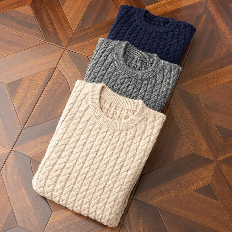 Three folded cable knit sweaters in cream, gray, and navy with ribbed cuffs are neatly stacked on patterned wooden floor tiles.