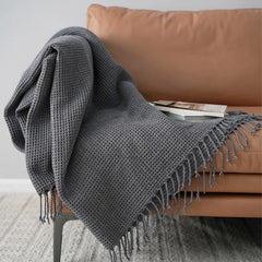 A gray knit waffle blanket with fringed edges is draped over a tan sofa, serving as a cozy decorative accent blanket. A book and magazine rest on the throw, creating an inviting scene against a light wall and patterned rug.
