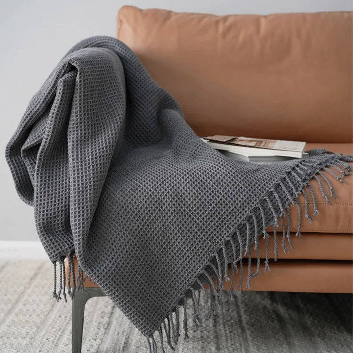 A gray knit waffle blanket with fringed edges is draped over a tan sofa, serving as a cozy decorative accent blanket. A book and magazine rest on the throw, creating an inviting scene against a light wall and patterned rug.