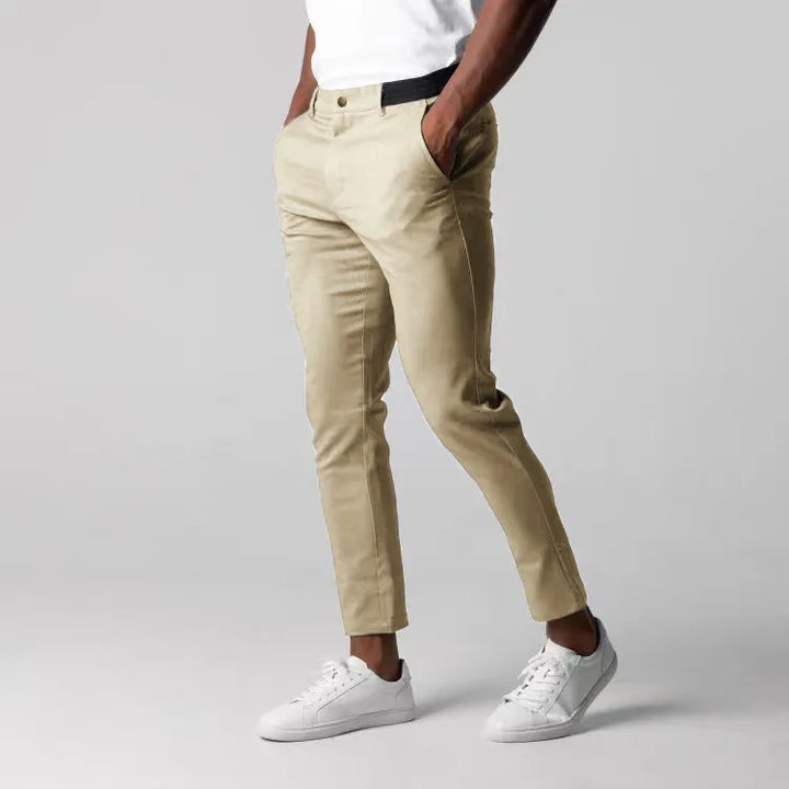 A person wearing beige slim-fit, ankle length pants with 4 way stretch, a white shirt, and white sneakers stands with hands in pockets against a plain gray background.