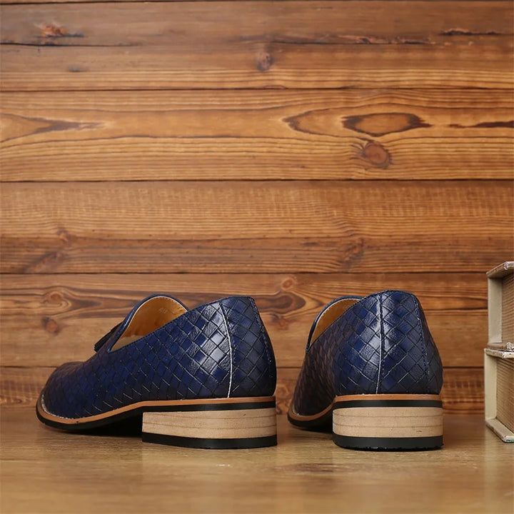 A pair of blue luxury footwear with a basketweave pattern and light brown soles, crafted from real leather, displayed on a wooden surface with a wood-paneled background. The shoes are positioned with the heels facing forward.