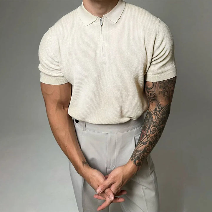 A man in a cream-colored quarter zip polo with ribbed detailing and light trousers stands with hands clasped in front. One arm is heavily tattooed. His head is cropped out of the frame.