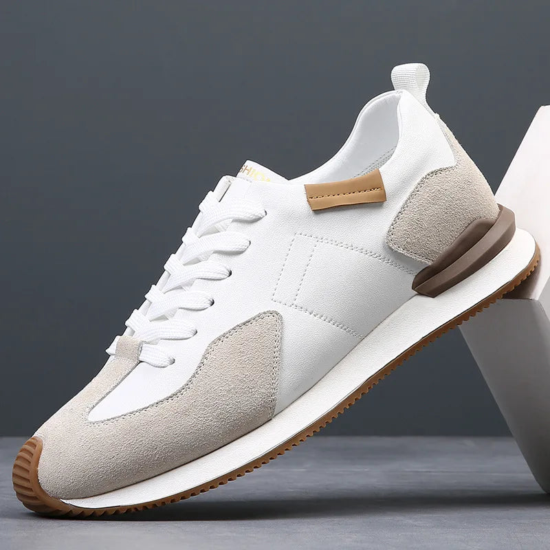 A modern white sneaker with beige suede accents, white laces, top-grain leather details, a brown heel tab, and a brown rubber sole—displayed against a gray background with geometric props.