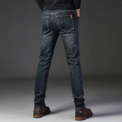 A person stands facing away, wearing slim-fit jeans made from durable fabric, brown shoes, and a black top. Their hands are in their pockets against a plain gray background.