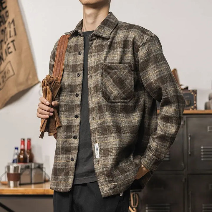 A person wearing a brown and gray plaid relaxed fit button-up shirt stands indoors with one hand in their pocket, holding a brown bag strap over their shoulder. The background shows bottles, lockers, and various items on a table.
