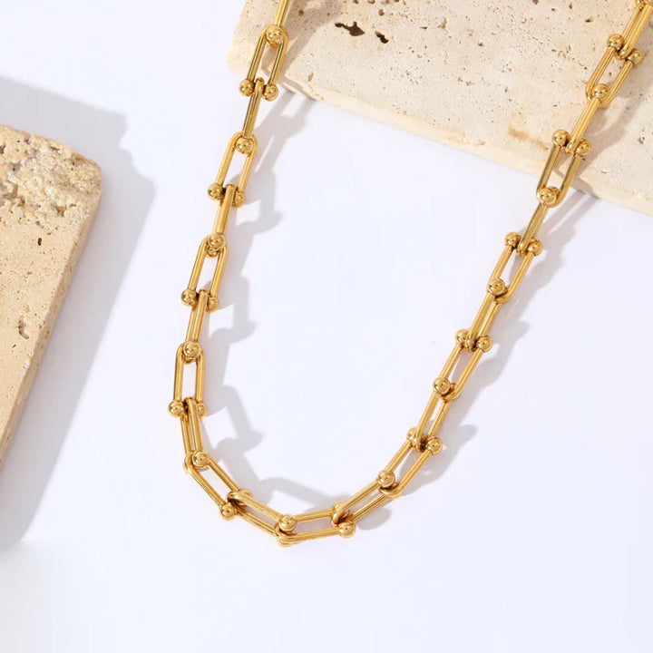 A gold statement chain necklace with elongated links and small spherical beads, arranged on a white surface with beige stone tiles, features an adjustable clasp for a perfect fit.