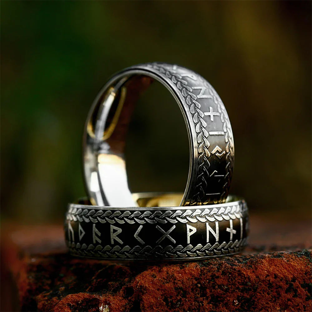 Two stainless steel Viking Rune Rings with intricate patterns rest on a reddish-brown surface, their engraved runic symbols glinting softly against a blurred green and dark background.