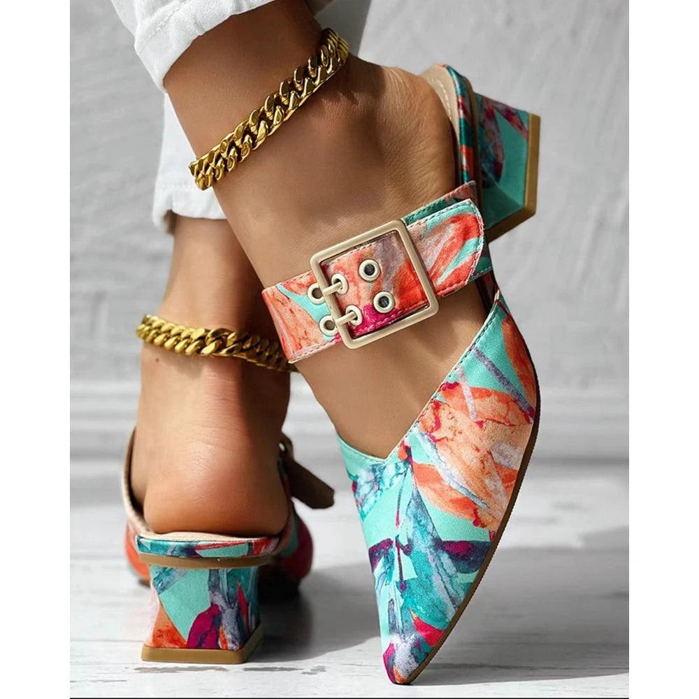 A close-up of colorful, floral-patterned summer mules with a chunky square heel and large buckle strap. The wearer also has a gold chain anklet and is dressed in white pants.
