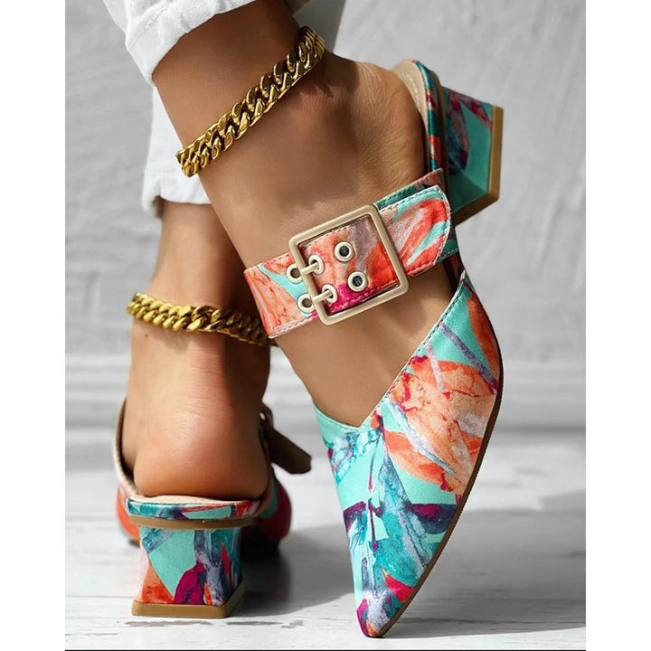 A close-up of colorful, floral-patterned summer mules with a chunky square heel and large buckle strap. The wearer also has a gold chain anklet and is dressed in white pants.