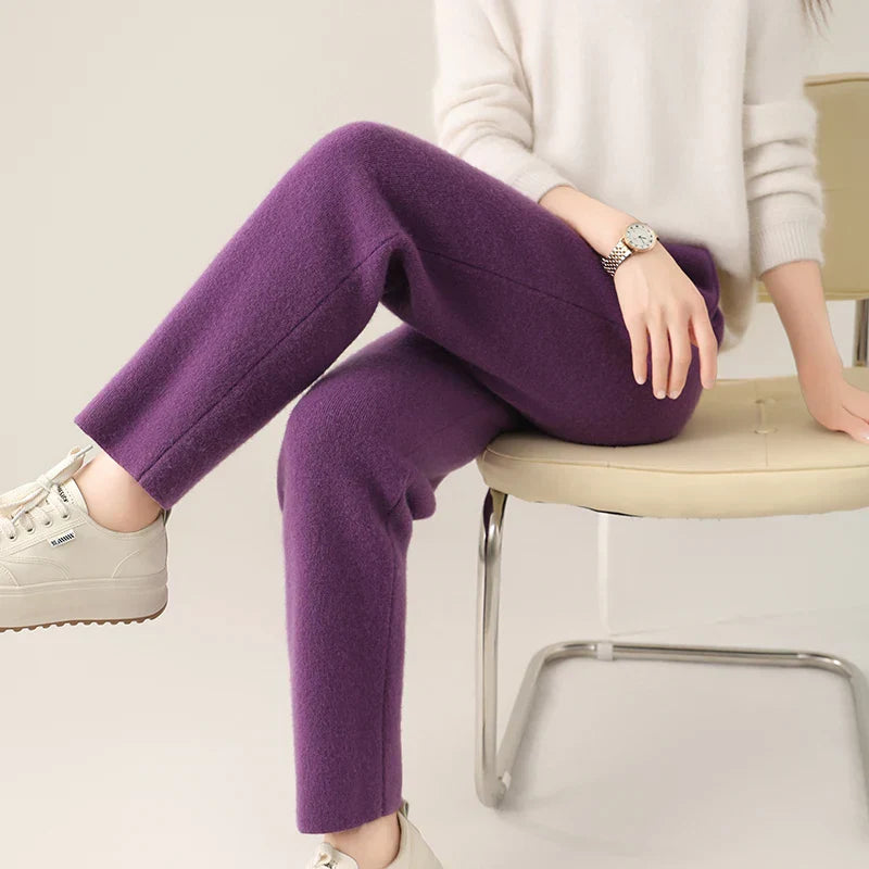 A person wearing a white sweater and purple pants with a slim tailored leg sits on a beige chair. Only the lower half of their body and left arm are visible. They are wearing white sneakers, a watch, and exude effortless style.