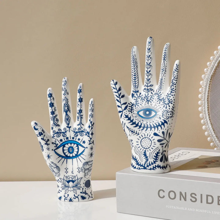 Two white ceramic hand sculptures with intricate blue eye and floral designs serve as unique ceramic jewelry stands, adding a touch of evil eye decor to a beige, minimalistic setting. One rests atop a book for boho-chic home décor flair.