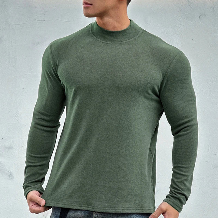 A man wearing a close-fitting, long-sleeve, olive green crewneck shirt made from a premium cotton blend and blue jeans stands against a plain, light gray background. His face is partially out of frame.