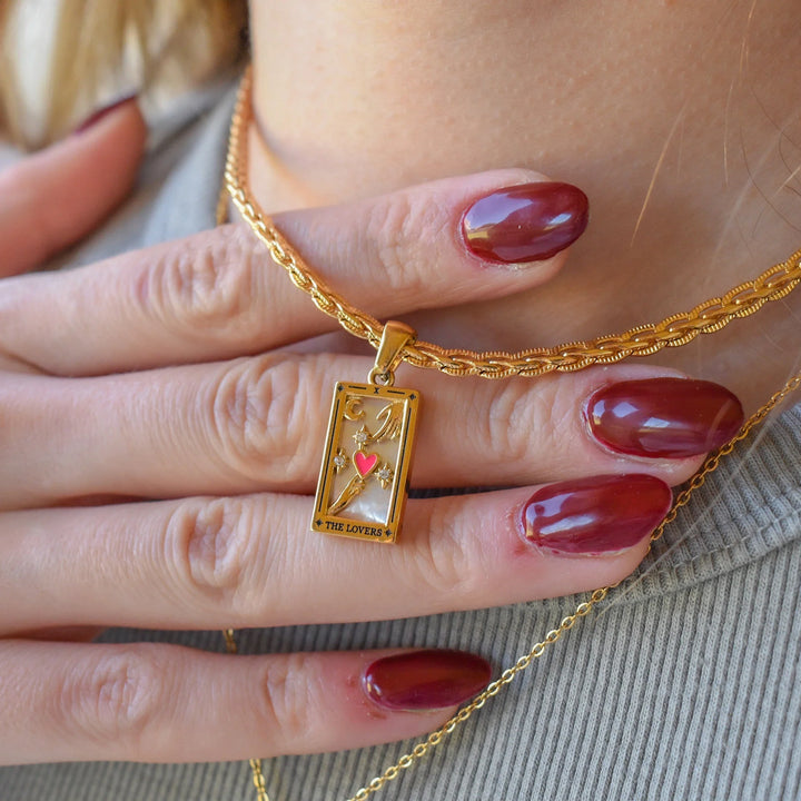 A hand with glossy dark red nails rests on a chest, displaying an 18K gold-plated rectangular pendant featuring a heart and the words THE LOVERS. Another gold chain necklace, possibly the Solaris Pendant Necklace, is also visible.