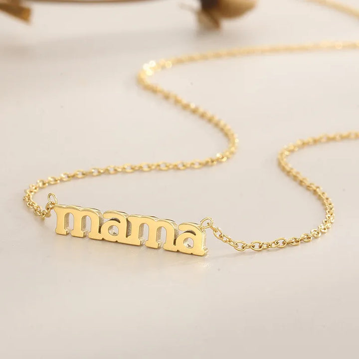A close-up of a gold Mama Forever Necklace with a pendant spelling mama in lowercase letters, resting on a smooth, light-colored surface.