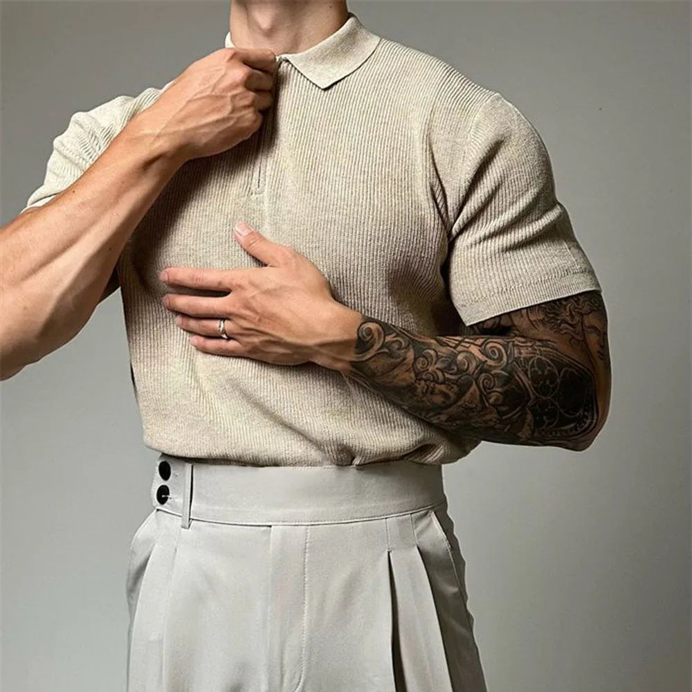 A person in a beige viscose polo with ribbed detailing and high-waisted light trousers adjusts their collar. One arm features a detailed black tattoo sleeve, and a ring is visible on their left hand. The background is plain and neutral.