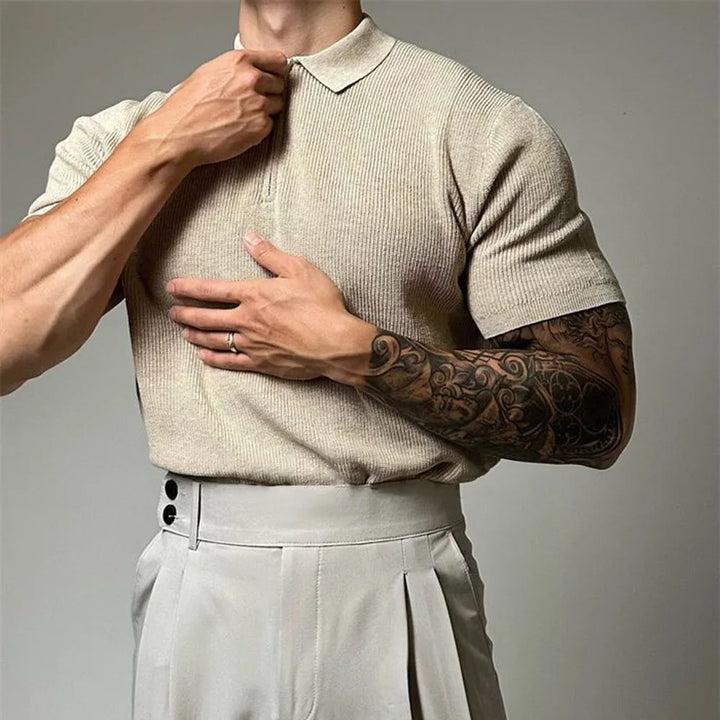 A person in a beige viscose polo with ribbed detailing and high-waisted light trousers adjusts their collar. One arm features a detailed black tattoo sleeve, and a ring is visible on their left hand. The background is plain and neutral.