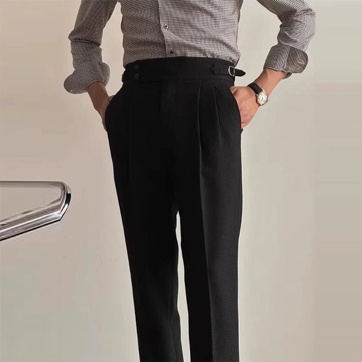 A person wearing a checkered long-sleeve shirt tucked into high-waisted black Havana Fit Slacks stands with hands in pockets. Part of a chair is visible on the left. The background is plain and light-colored.