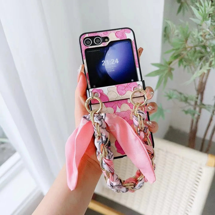 A hand holds a pink floral foldable smartphone with slim design, featuring a matching braided wrist strap and scarf, displaying the time 20:24 on the small external screen. A leafy plant and a white chair are in the background.