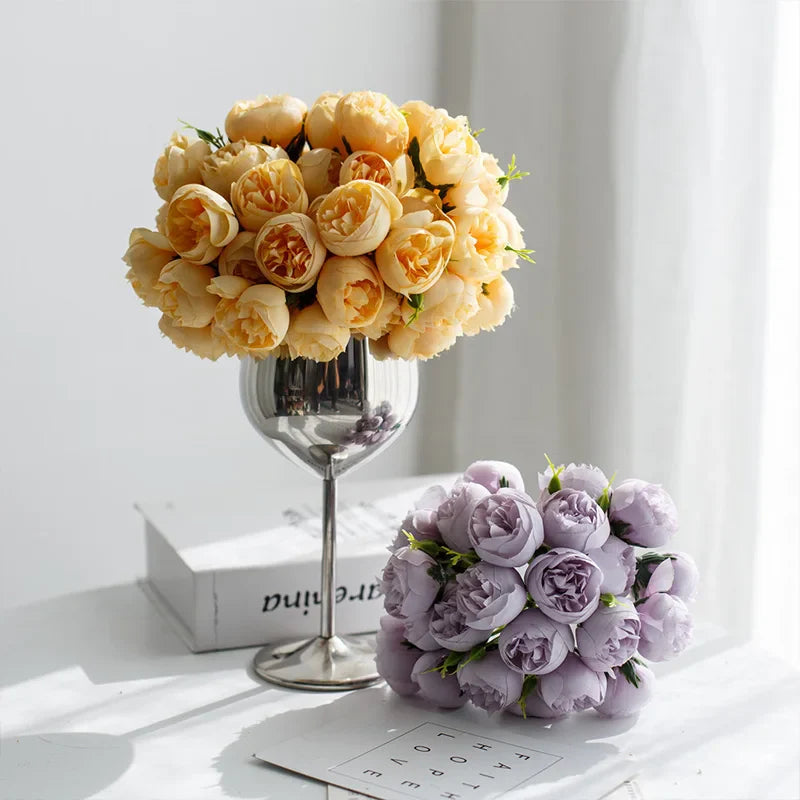 A bouquet of yellow roses with faux petals in a wine glass vase and a bouquet of light purple roses resting on a magazine, both displayed on a sunlit white surface near a window.