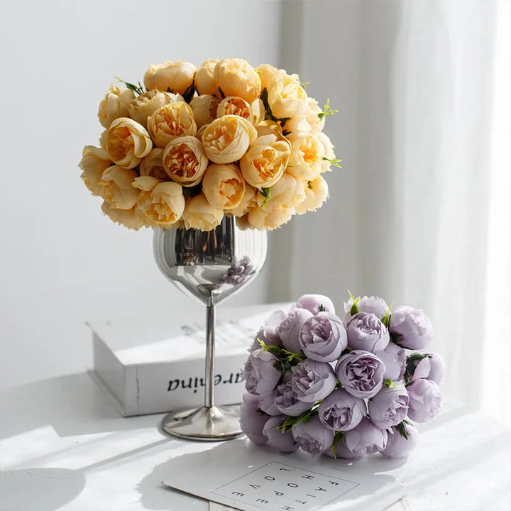 A bouquet of yellow roses with faux petals in a wine glass vase and a bouquet of light purple roses resting on a magazine, both displayed on a sunlit white surface near a window.
