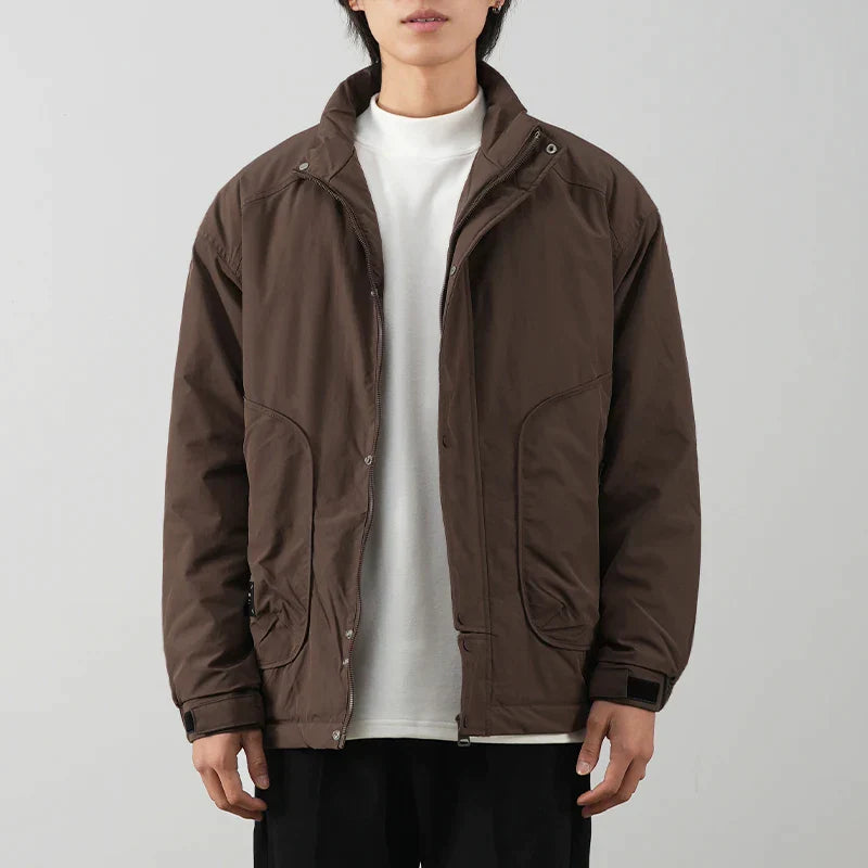 A person wearing a minimalist brown jacket with luxurious cotton lining over a white t-shirt and black pants stands against a plain light gray background. The jacket has a high collar and large front pockets.