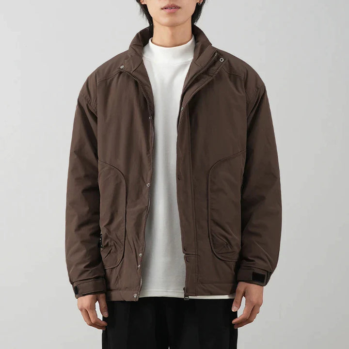 A person wearing a minimalist brown jacket with luxurious cotton lining over a white t-shirt and black pants stands against a plain light gray background. The jacket has a high collar and large front pockets.