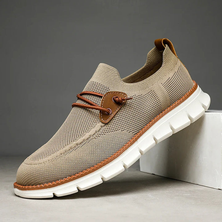 A beige casual sneaker with a breathable mesh upper, white thick rubber sole, brown trim, and decorative lace detail—these breathable loafers are displayed at an angle with the toe resting on a white block.