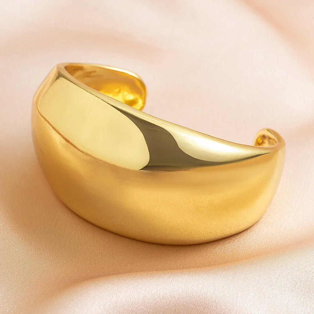 A wide, polished gold cuff bracelet with a smooth, shiny surface and slightly curved shape rests on soft light pink satin fabric—a striking piece of unisex jewelry for any collection.