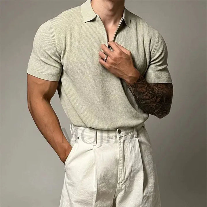A man wearing a light beige quarter zip polo with ribbed detailing and high-waisted white pants, one hand in his pocket and the other adjusting his collar; his right arm features a detailed tattoo sleeve.