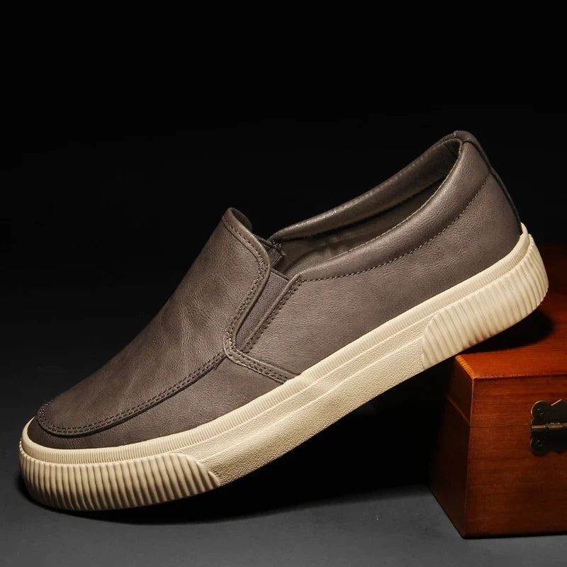 A men’s slip-on brown leather shoe with a leather insole and hybrid rubber sole is displayed propped up on a wooden box against a dark background.