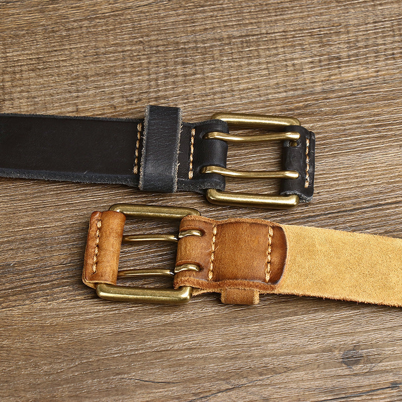 Two genuine leather belts with brass buckles lie parallel on a wooden surface, showcasing heritage craftsmanship. One is black with light stitching, the other tan with matching thread. Both boast a worn, rustic look.