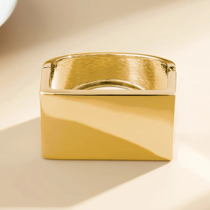 A rectangular, gold-colored metal ring with a smooth, polished surface and a slightly textured inner band, this unisex jewelry piece features a subtle geometric design, photographed on a neutral beige background.