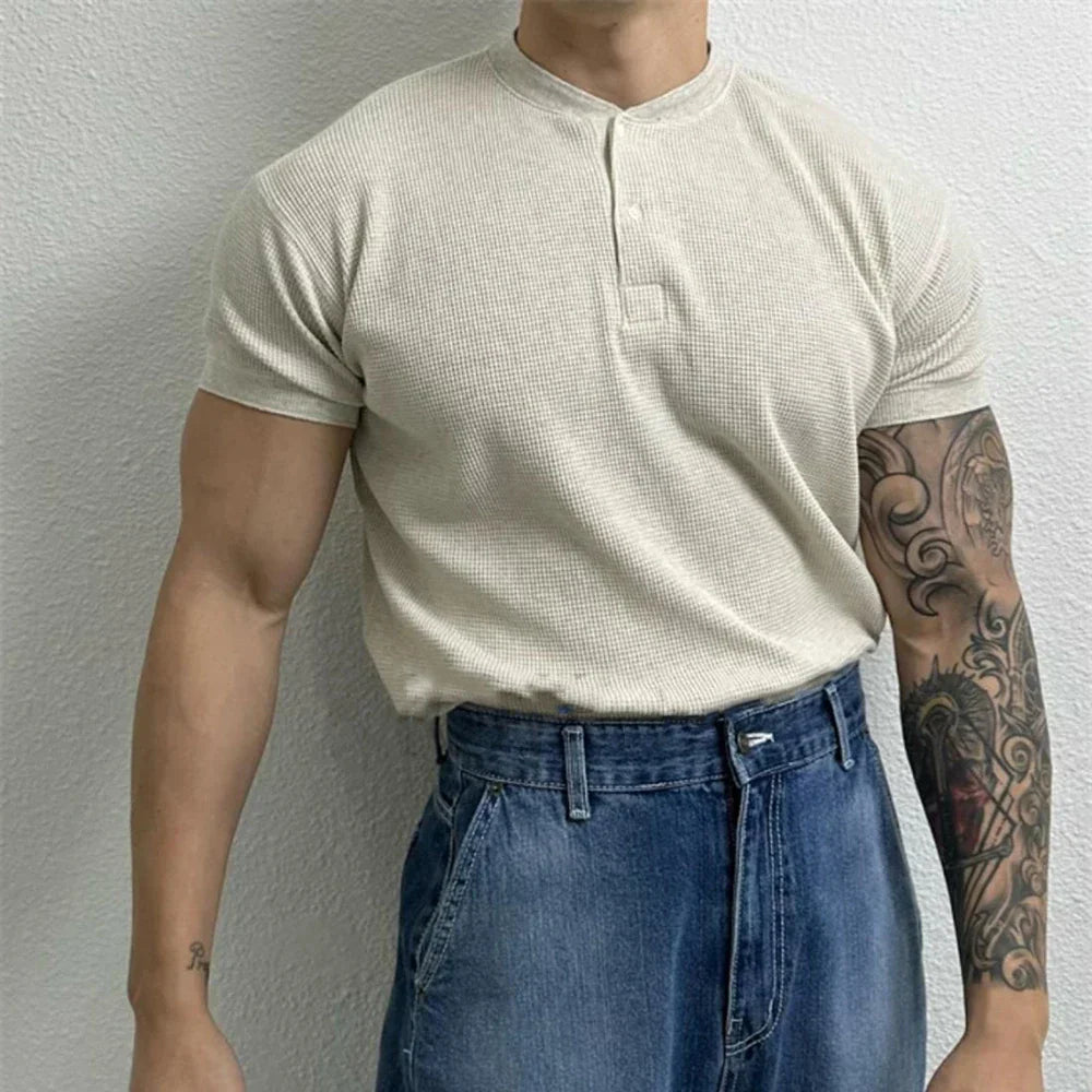 A person wearing a short-sleeve, beige ribbed texture shirt with a Henley neckline tucked into high-waisted blue jeans stands against a light-colored wall. Their right arm is tattooed, while their left arm is untattooed. Their face is not visible.