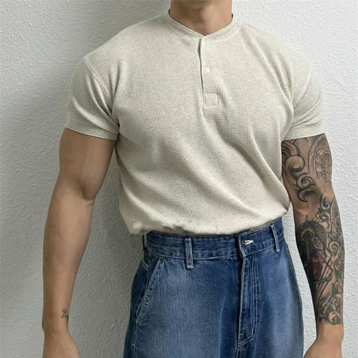 A person wearing a short-sleeve, beige ribbed texture shirt with a Henley neckline tucked into high-waisted blue jeans stands against a light-colored wall. Their right arm is tattooed, while their left arm is untattooed. Their face is not visible.